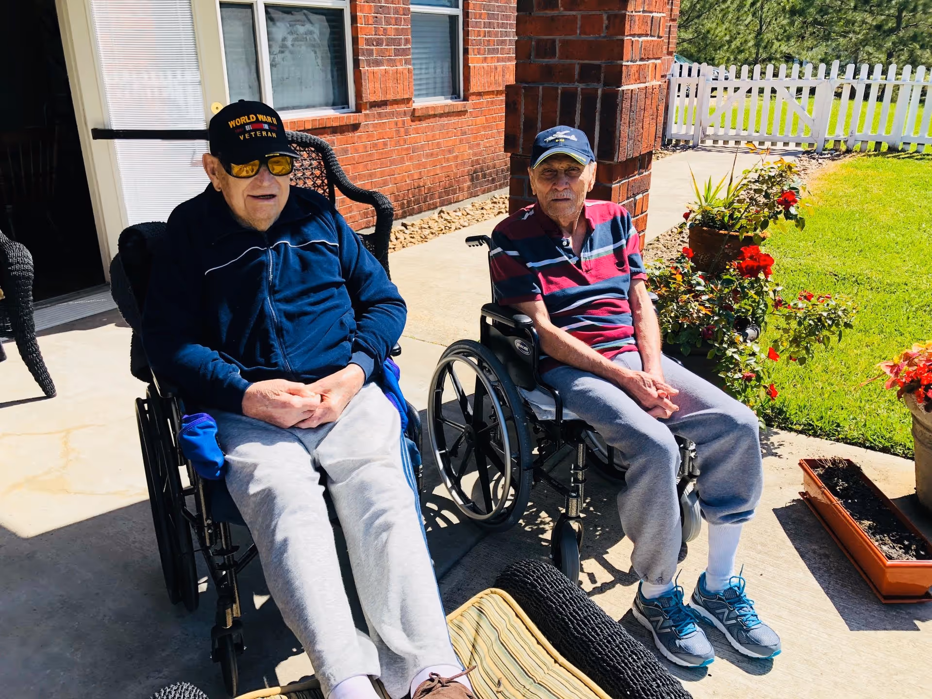 Two elderly men sitting in wheelchairs outside on a sunny patio next to a brick building. One man is wearing a black 'World War II Veteran' cap and sunglasses, while the other is wearing a navy blue cap and a striped polo shirt. There are flower pots with red flowers and a white picket fence in the background.