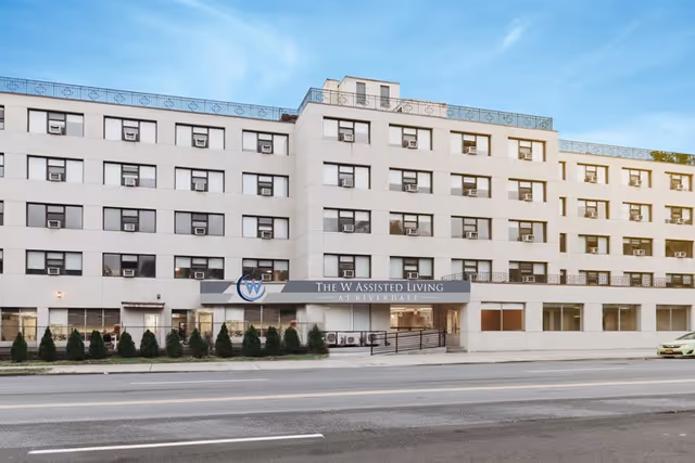 Front exterior of a multi-story assisted living building with a sign reading 'The W Assisted Living at Riverdale'.