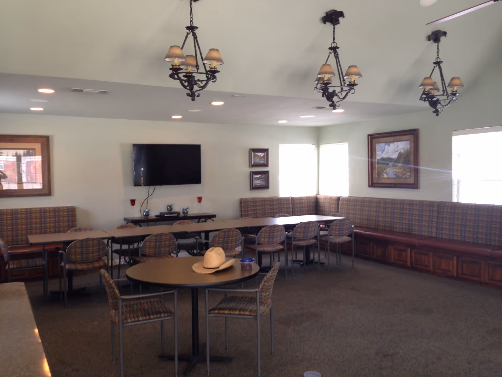 Spacious communal dining/activity room with round and rectangular tables, patterned chairs, built-in bench seating, a wall-mounted TV, and hanging chandeliers.