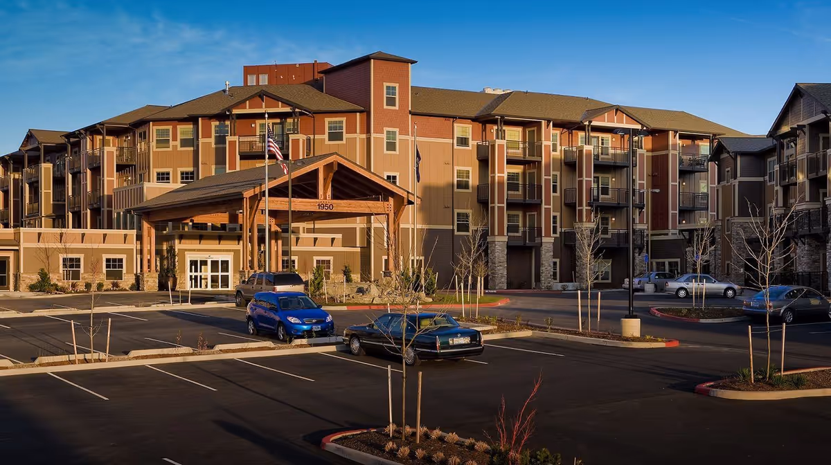 Exterior view of The Springs at Tanasbourne senior living facility showing a multi-story building with balconies, a covered entrance with wooden beams, several parked cars in the parking lot, and clear blue sky.