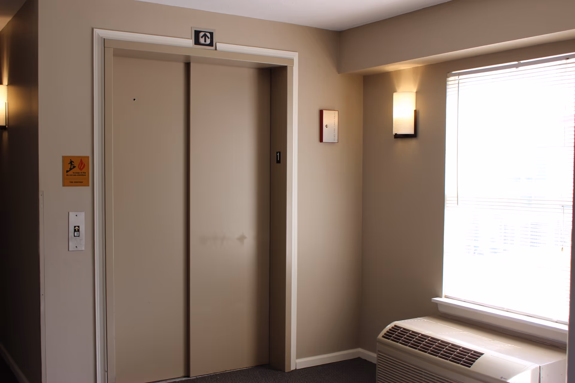 Elevator doors in a hallway with beige walls, a window with blinds, a wall-mounted light fixture, and a heating or air conditioning unit below the window.