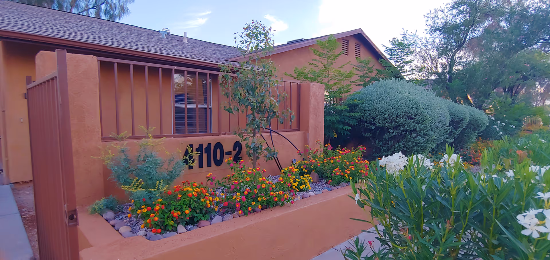 Exterior view of a building at Groves Assisted Living at Lemon Tree featuring a tan stucco wall with the number 110-2, surrounded by a garden with colorful flowers and green shrubs under a partly cloudy sky.