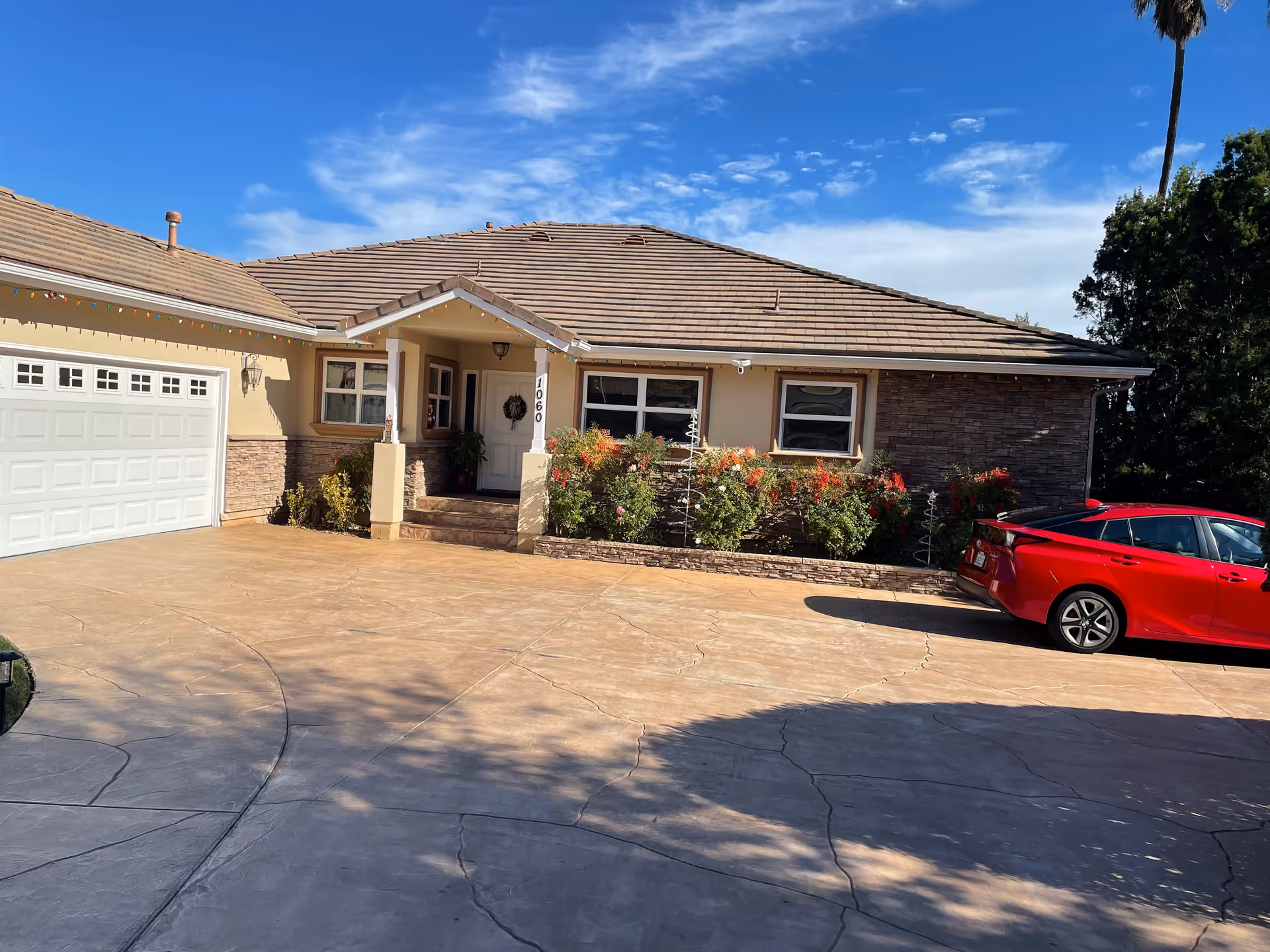 Front exterior view of a single-story assisted living facility with a tiled roof, beige walls, a white garage door, a small porch with steps, and a garden bed with flowering bushes. A red car is parked on the driveway under a clear blue sky.
