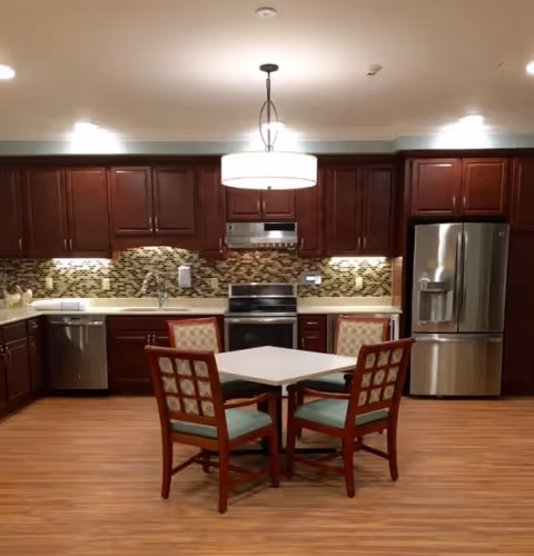 Well-lit communal kitchen with dark wooden cabinets, stainless steel appliances, tiled backsplash, and a small table with four chairs in the center.
