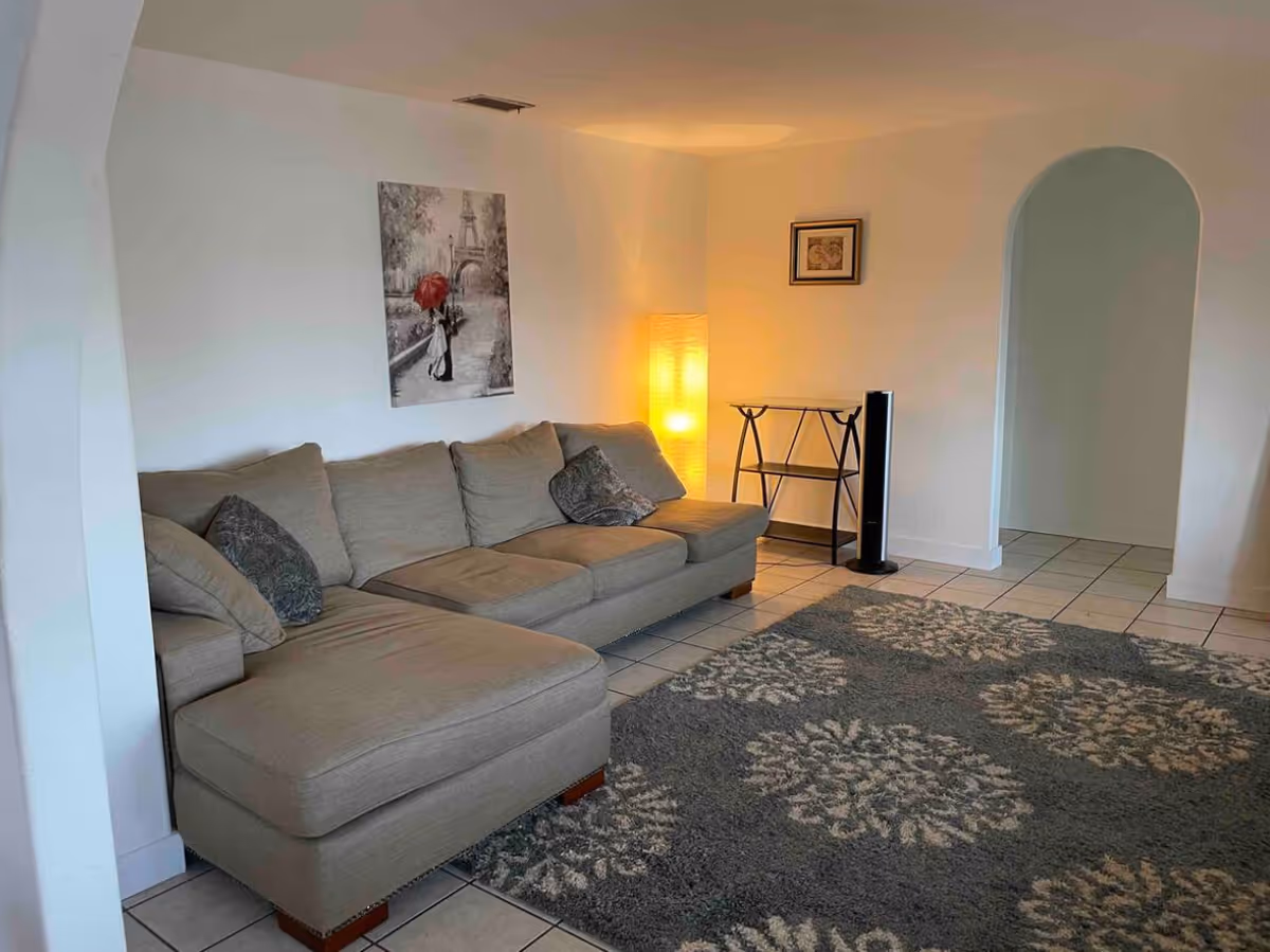 A cozy living room with a beige sectional sofa, a tall floor lamp emitting warm light, a small glass and metal table, a standing fan, and a large patterned area rug on tiled flooring. The walls are white with two framed pictures, one featuring a couple with a red umbrella near the Eiffel Tower.