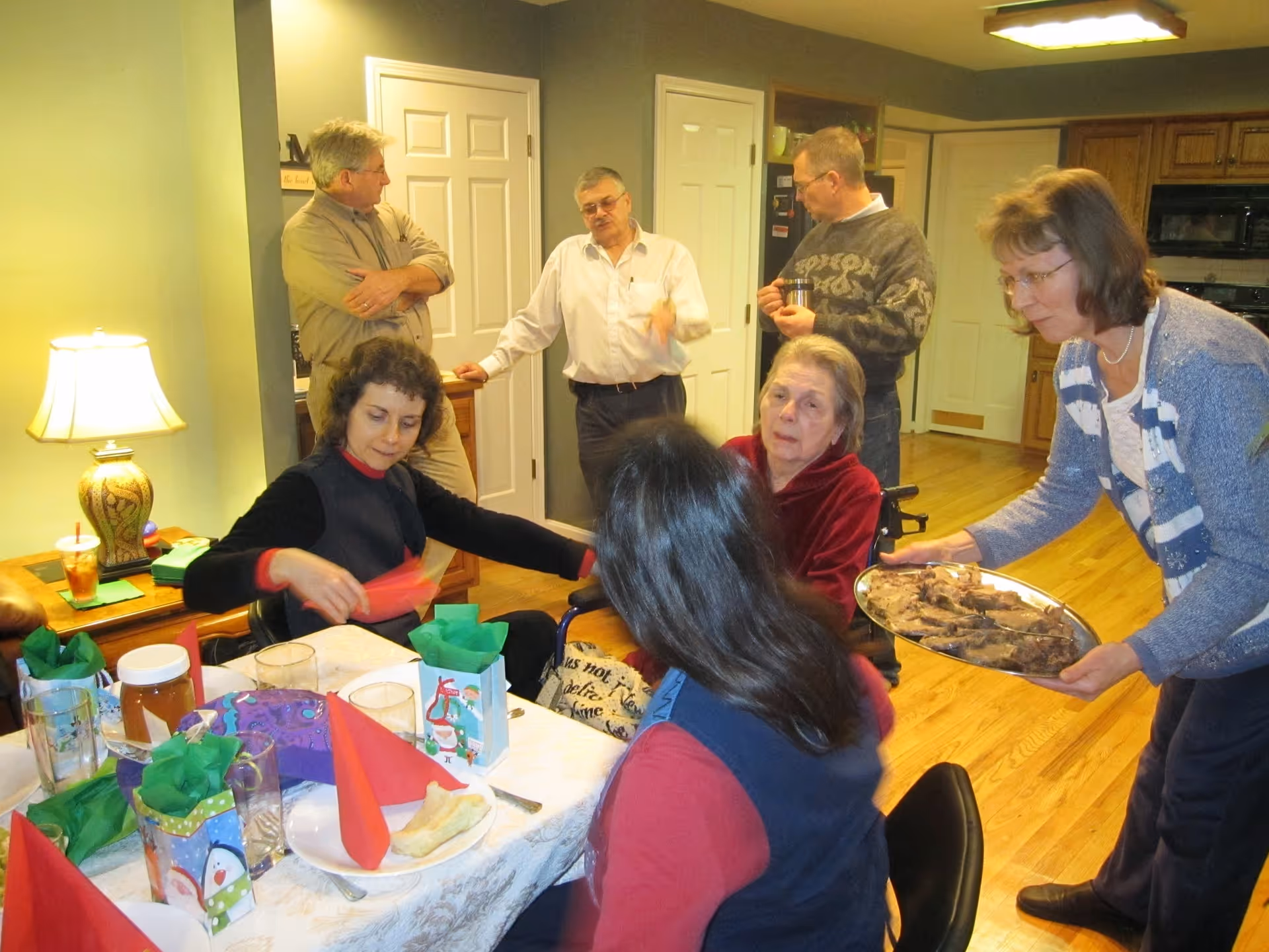 A group of six people gathered in a warmly lit dining area. Three women are seated around a table set with holiday-themed gift bags, red napkins, and glasses. One woman is serving a tray of food to an elderly woman in a wheelchair. Three men stand in the background engaged in conversation. The room has wooden floors and kitchen cabinets visible in the background.