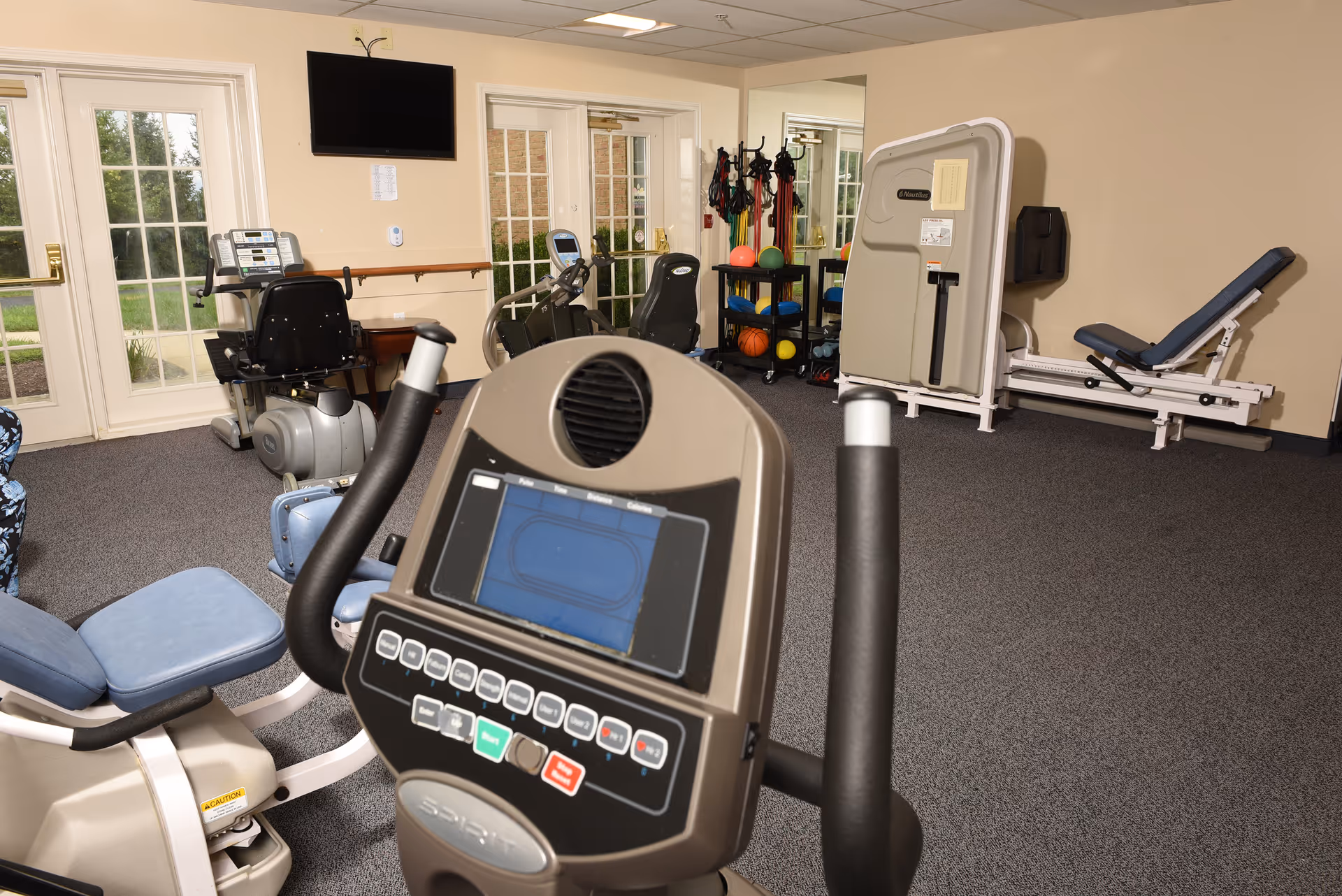 Interior view of a fitness room in a senior living facility with exercise equipment including stationary bikes, a weight machine, resistance bands, and exercise balls. There are large windows and glass doors letting in natural light, and a wall-mounted TV is visible.