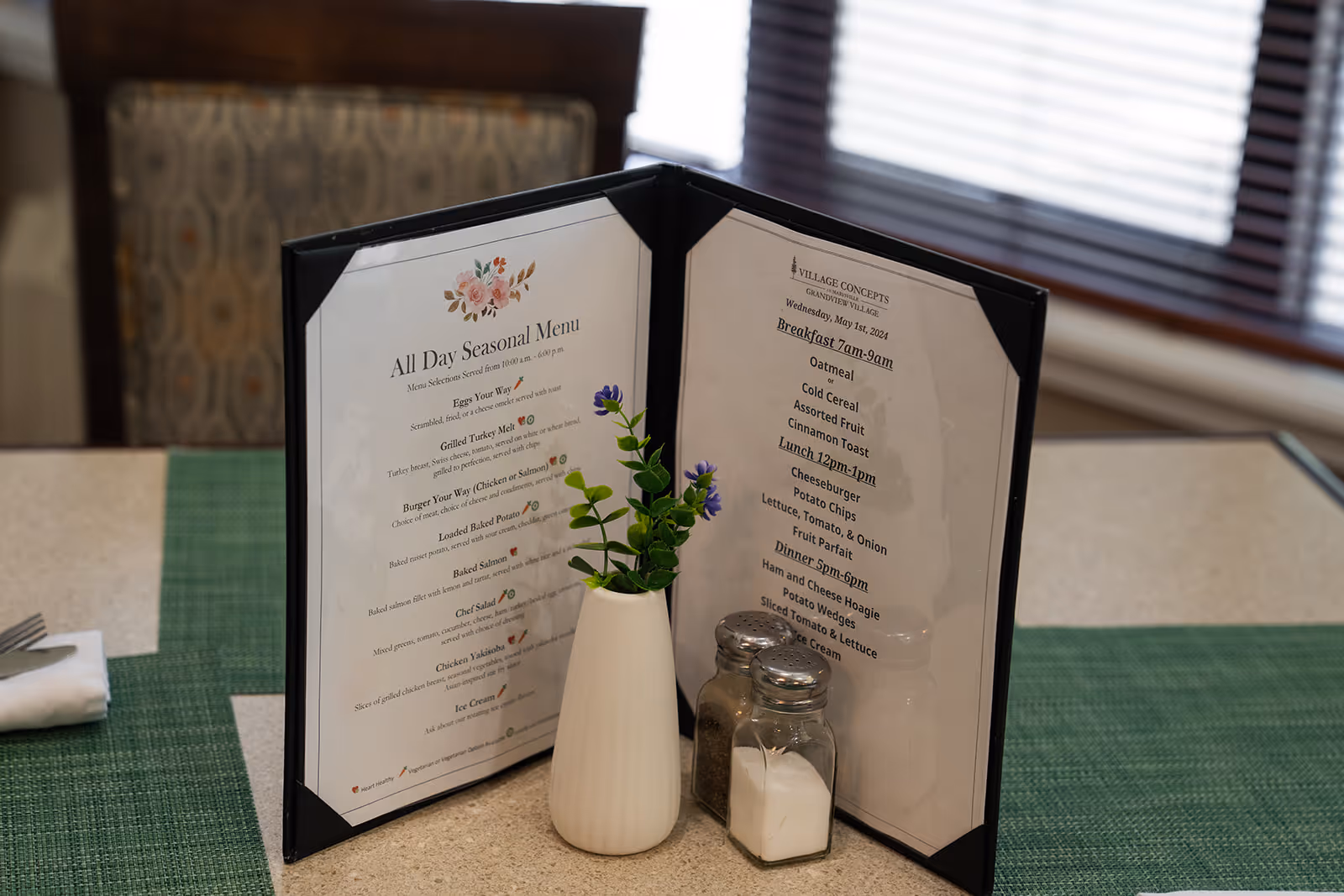 A dining table with a green placemat, a folded menu in a black holder displaying an all day seasonal menu and daily meal times, a small white vase with purple flowers, and salt and pepper shakers. The background shows a cushioned chair and window blinds.