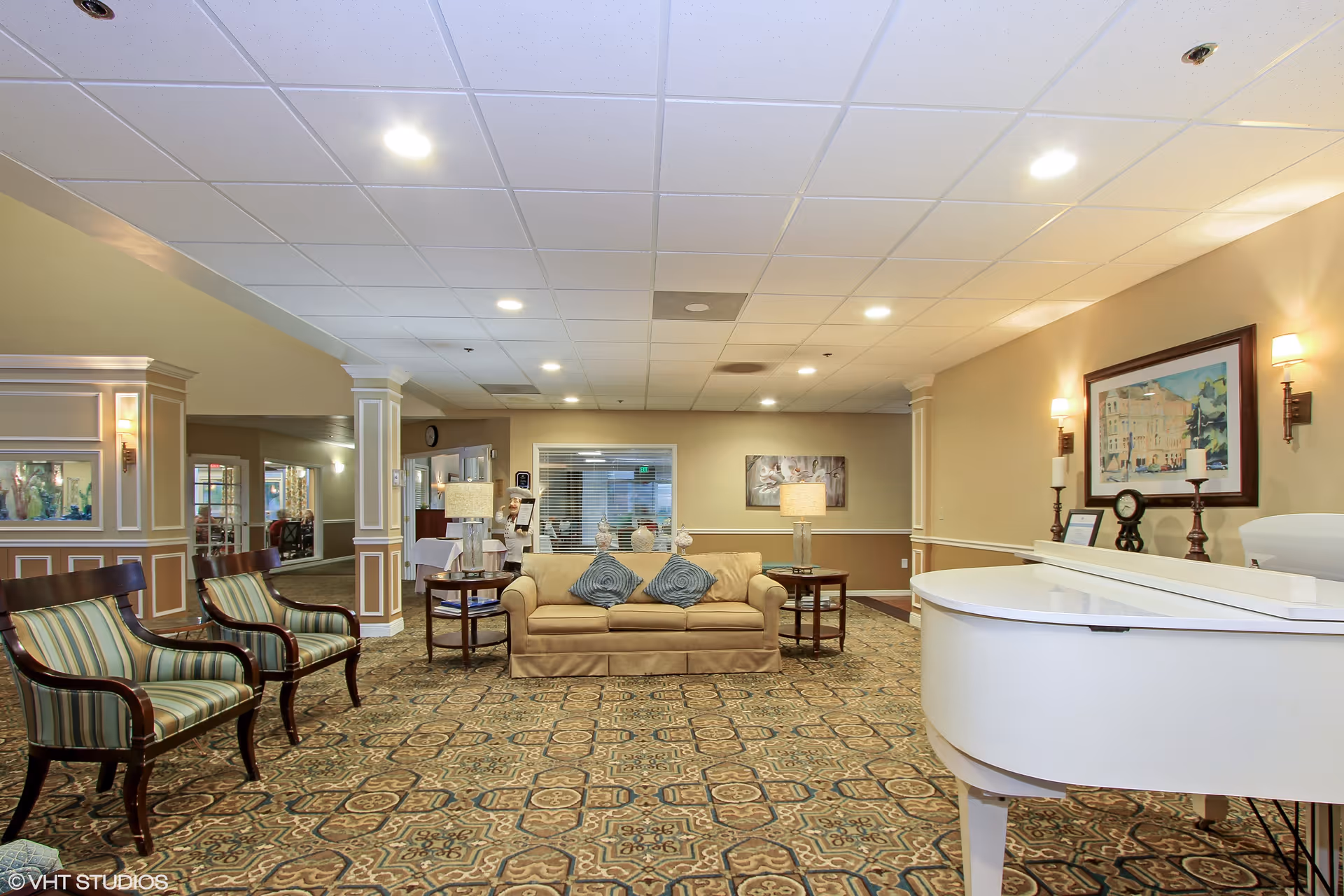 A spacious and well-lit common area in a senior living facility featuring a beige sofa with two blue patterned pillows, two striped armchairs, a white grand piano, and side tables with lamps. The room has patterned carpet, beige walls with white trim, framed artwork, and recessed ceiling lights.