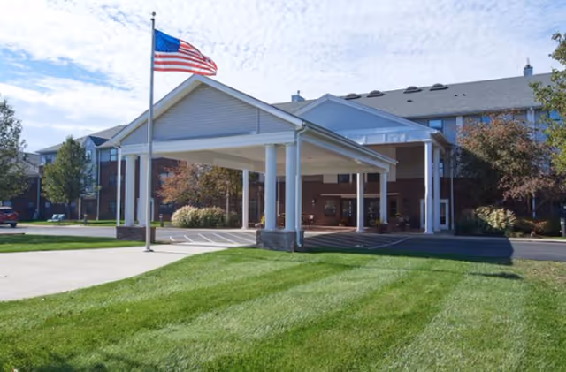 Front exterior view of a senior living facility with a covered entrance supported by white columns, an American flag on a flagpole, green lawn, and trees under a partly cloudy sky.