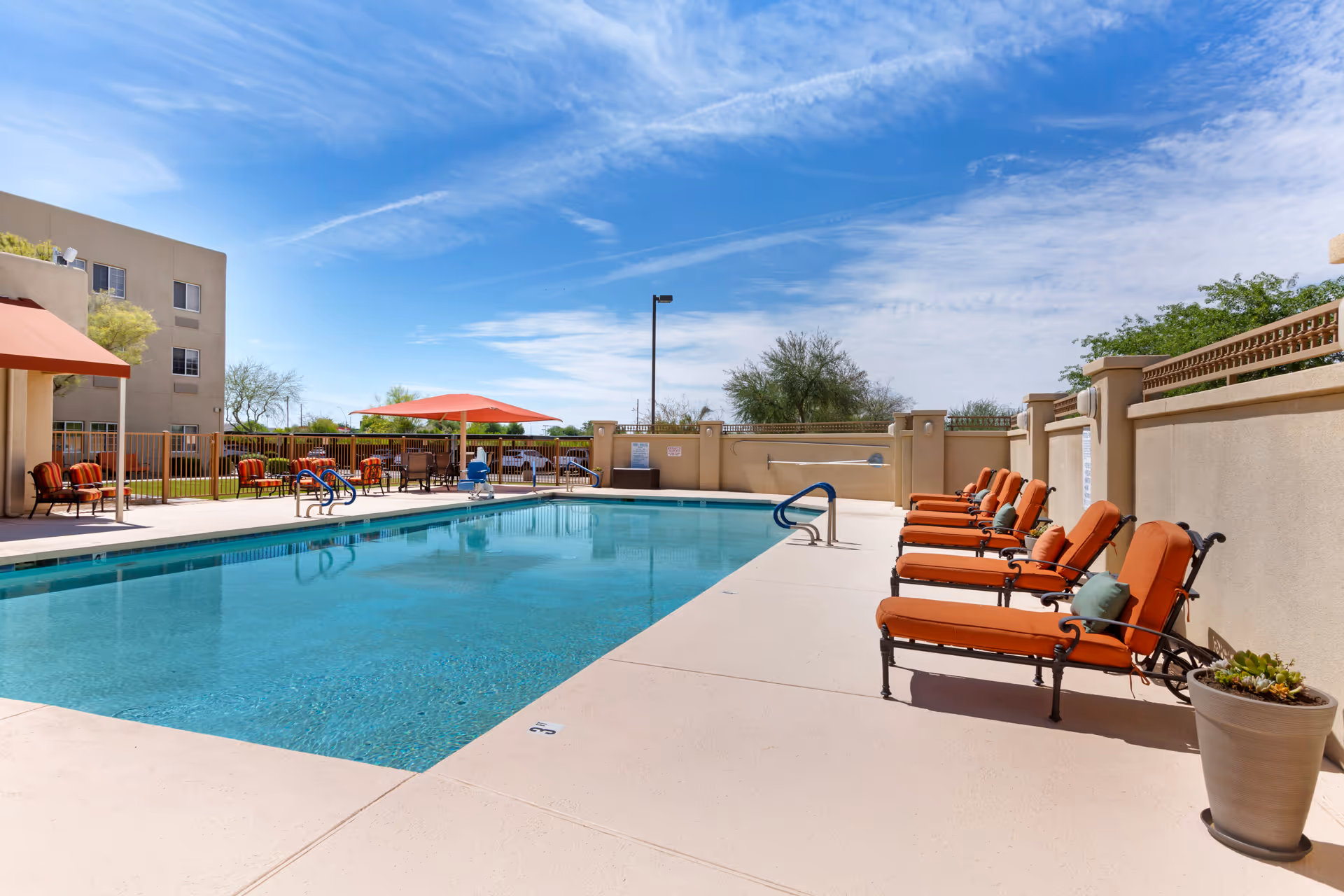 Outdoor swimming pool with orange-cushioned lounge chairs and umbrellas beside a beige building under a blue sky.