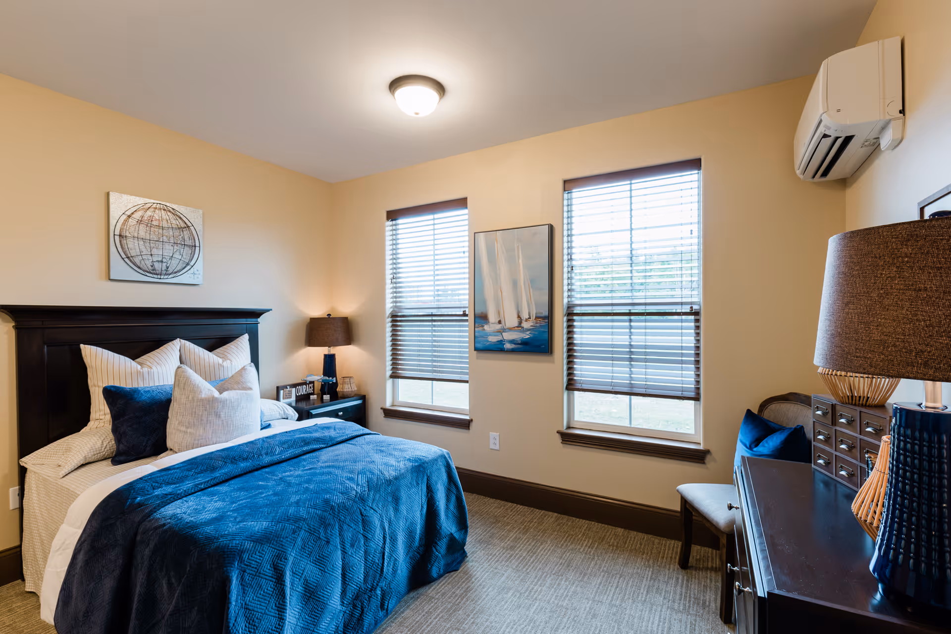 A cozy bedroom with a neatly made bed featuring blue and white bedding and multiple pillows. The room has beige walls, two windows with blinds, a dark wooden headboard, a nightstand with a lamp, and a dresser with another lamp and a chair beside it. There are two pieces of wall art, one above the bed and one between the windows.