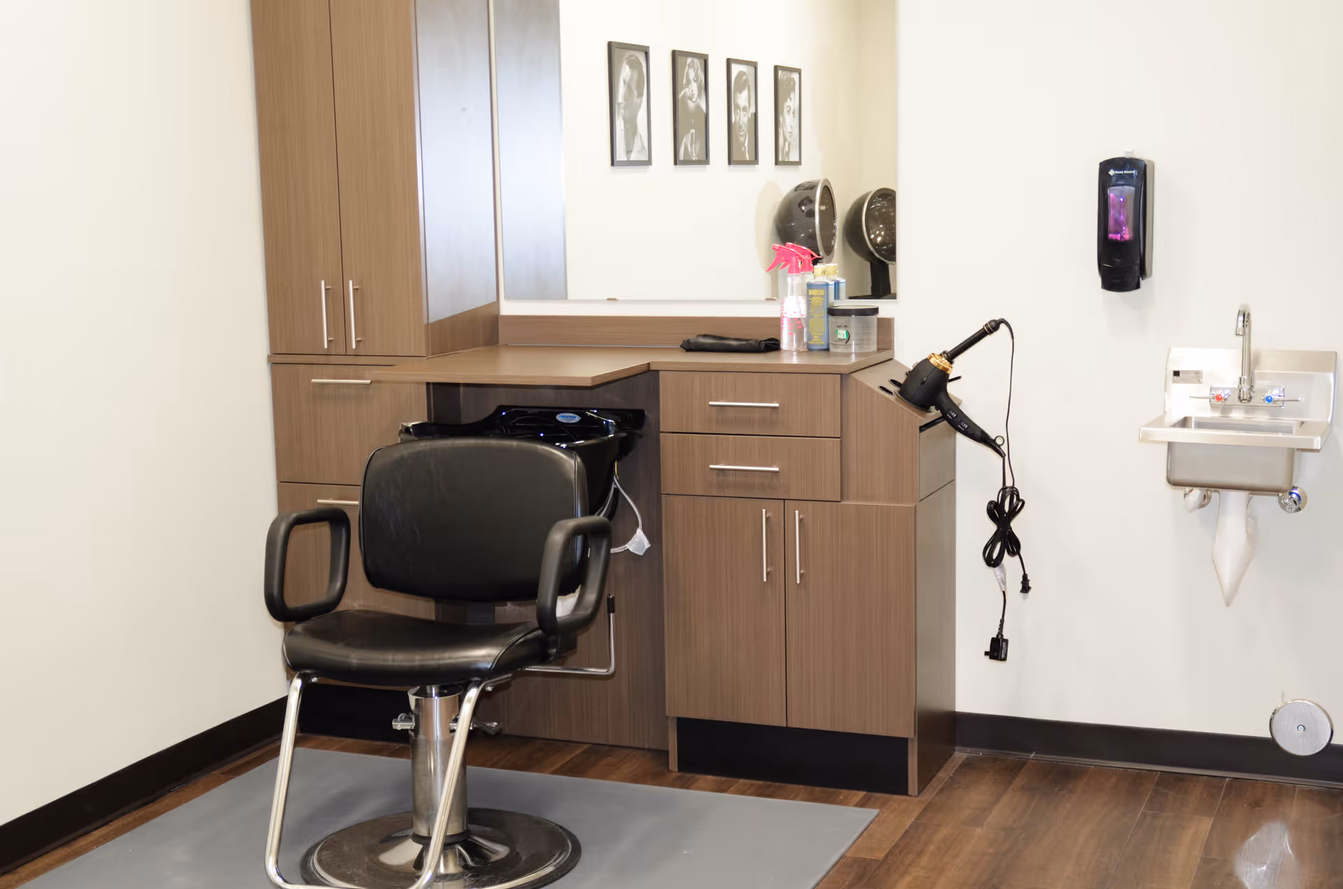 Interior view of a hair salon area in a senior living facility featuring a black salon chair in front of a wooden cabinet with a mirror, hair dryer, and hair washing station. A small wall-mounted sink and soap dispenser are visible on the right side.