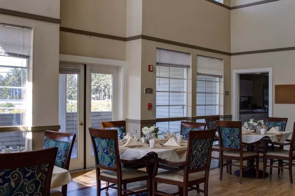 Bright dining room with tables set for meals and patterned wooden chairs in an assisted living facility.