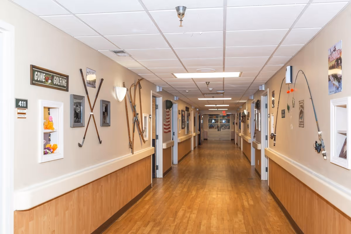A long hallway in a senior living facility with wooden flooring and beige walls decorated with vintage sports equipment including golf clubs, skis, and fishing rods. Several framed pictures and signs, including one that says 'Gone Golfing,' are mounted on the walls. Room doors line both sides of the hallway, and ceiling lights illuminate the space.