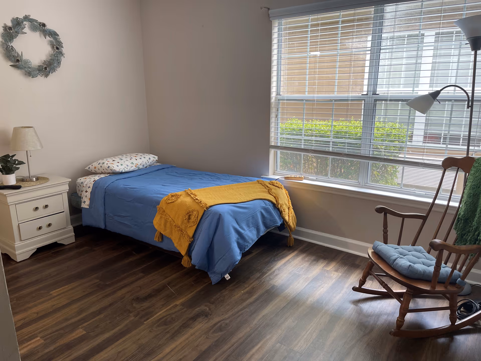 Simple furnished bedroom with a single bed dressed in blue bedding and a yellow throw, a nightstand, large window, and a wooden rocking chair.
