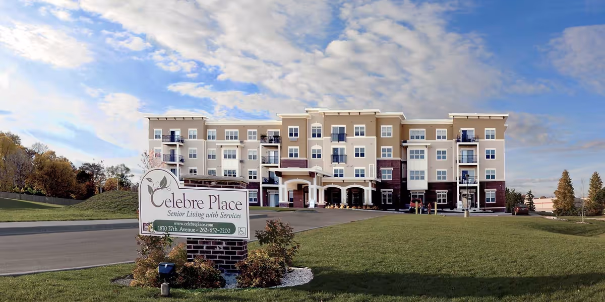 Exterior view of Celebre Place, a senior living facility with a multi-story building featuring balconies and a covered entrance. The foreground shows a green lawn and a sign with the facility's name, website, address, and phone number. The sky is partly cloudy with patches of blue.