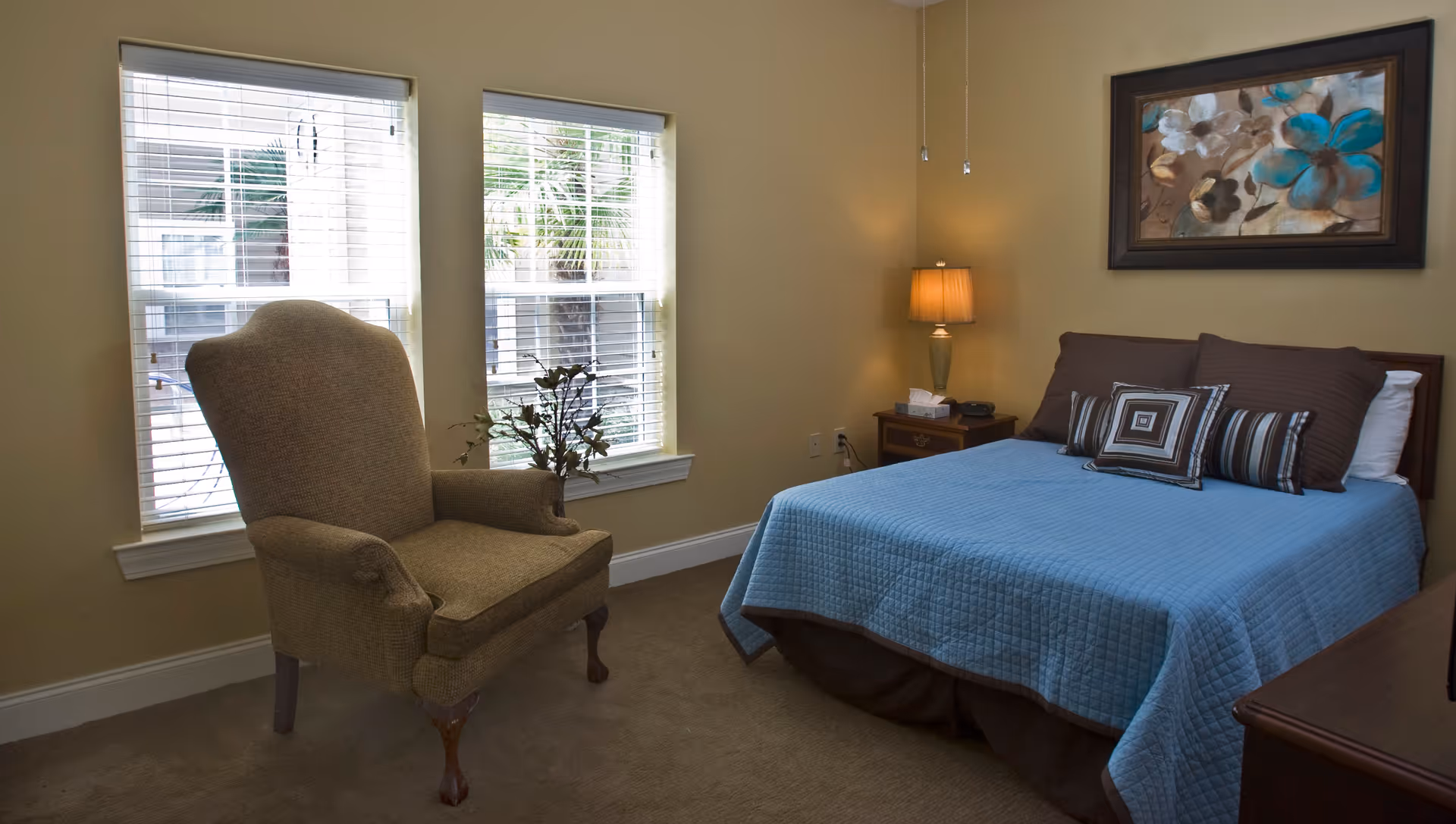 A cozy bedroom with a bed covered in a blue quilt and several decorative pillows. There is a beige upholstered armchair near two windows with white blinds. A wooden nightstand with a lamp and a tissue box is next to the bed. A framed floral painting hangs on the wall above the bed.