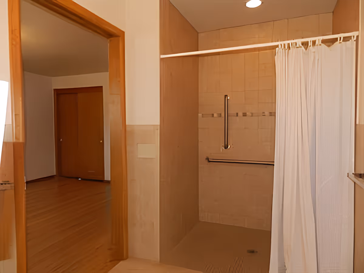Interior view of a bathroom with a walk-in shower featuring beige tiles, a white shower curtain, and metal grab bars. The bathroom opens into a room with wooden flooring and a closed wooden door.