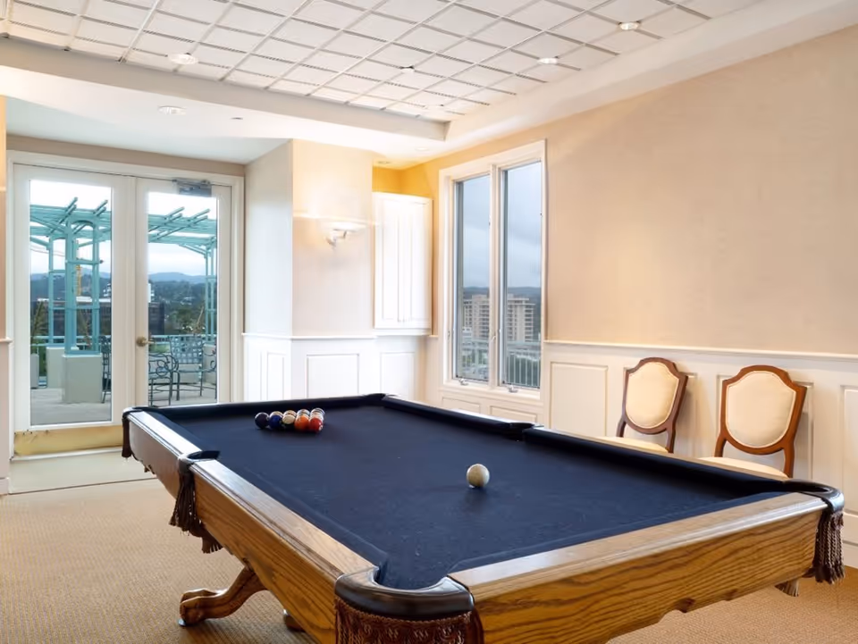 A bright interior recreation room featuring a pool table with balls, two chairs, and glass doors opening to an outdoor terrace.