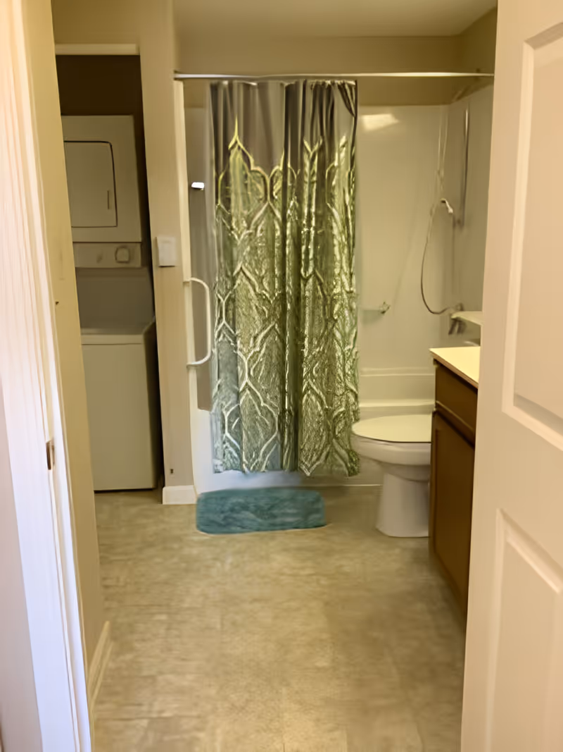 Bathroom with a shower/tub behind a green patterned curtain, a toilet and vanity to the right, and a stacked washer-dryer in a closet to the left.
