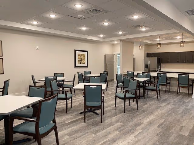 A spacious dining area with multiple square tables and teal cushioned chairs arranged neatly on a wood-patterned floor. The room has a modern ceiling with recessed lighting and pendant lights hanging over a counter area with cabinets and a refrigerator in the background. A framed artwork is visible on the wall.