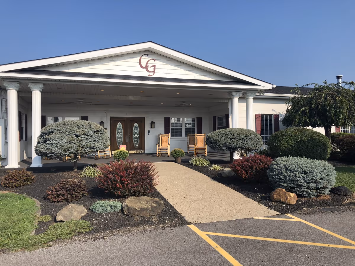 Front exterior view of a single-story building with white columns and a covered entrance. The building has a sign with the letters 'CG' above the entrance. There are shrubs, small trees, and rocks lining the walkway leading to double wooden doors with glass panels. Two wooden rocking chairs are placed on the porch.