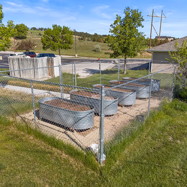 Outdoor fenced garden area with multiple raised metal garden beds filled with soil, surrounded by grass and trees, with a parking lot and building visible in the background under a clear blue sky.