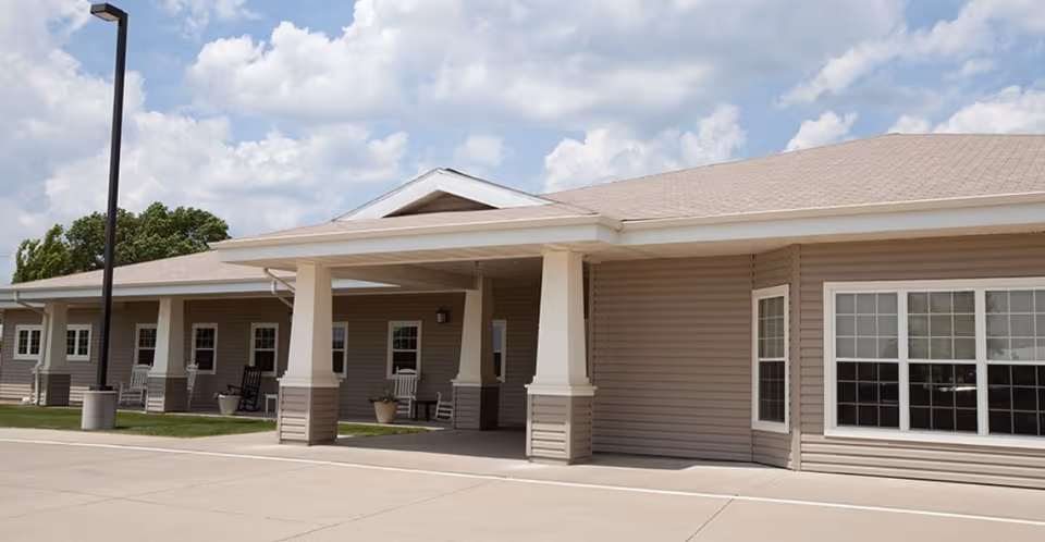 Front exterior of a single-story beige assisted living building with a covered porte-cochere, columns, windows, and rocking chairs.