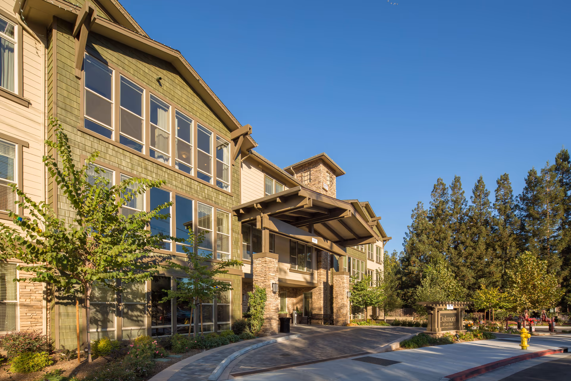 Exterior view of a multi-story senior living facility building with green and beige siding, large windows, a covered entrance, landscaped trees and shrubs, and a clear blue sky.