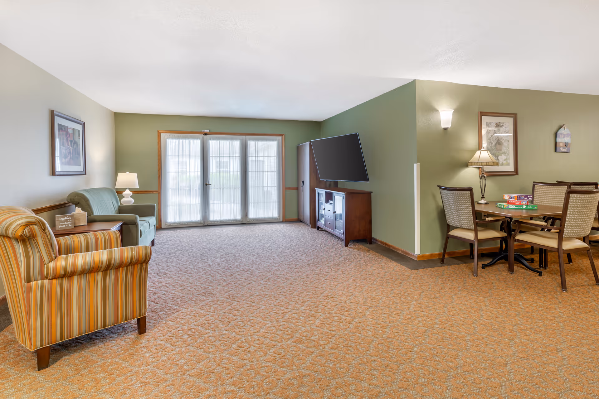 A senior living facility common area with a striped armchair and a green armchair near a side table with a lamp and a decorative sign. There is a large window with sheer curtains letting in natural light. A flat-screen TV is mounted on a green wall above a wooden cabinet. To the right, there is a dining table with four chairs and board games on top, along with a lamp and framed artwork on the wall.