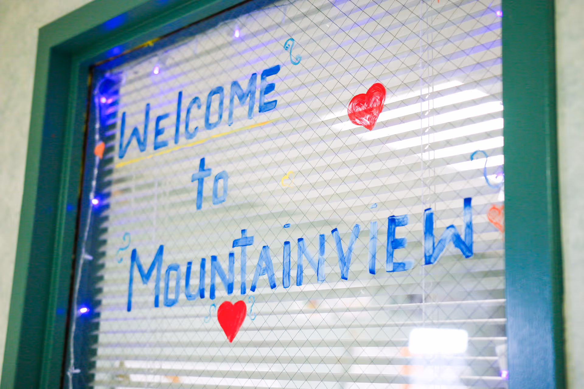 A glass window with a green frame displaying a handwritten message in blue and red that says 'Welcome to Mountainview' with red hearts and decorative swirls around the text. The window has a wire mesh pattern and blinds behind it.