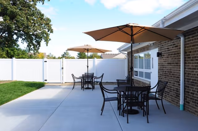 Outdoor patio area with two round tables, each with four black metal chairs and a large beige umbrella. The patio is adjacent to a brick building with a white fence enclosing the area and green grass visible on the left side under a partly cloudy sky.