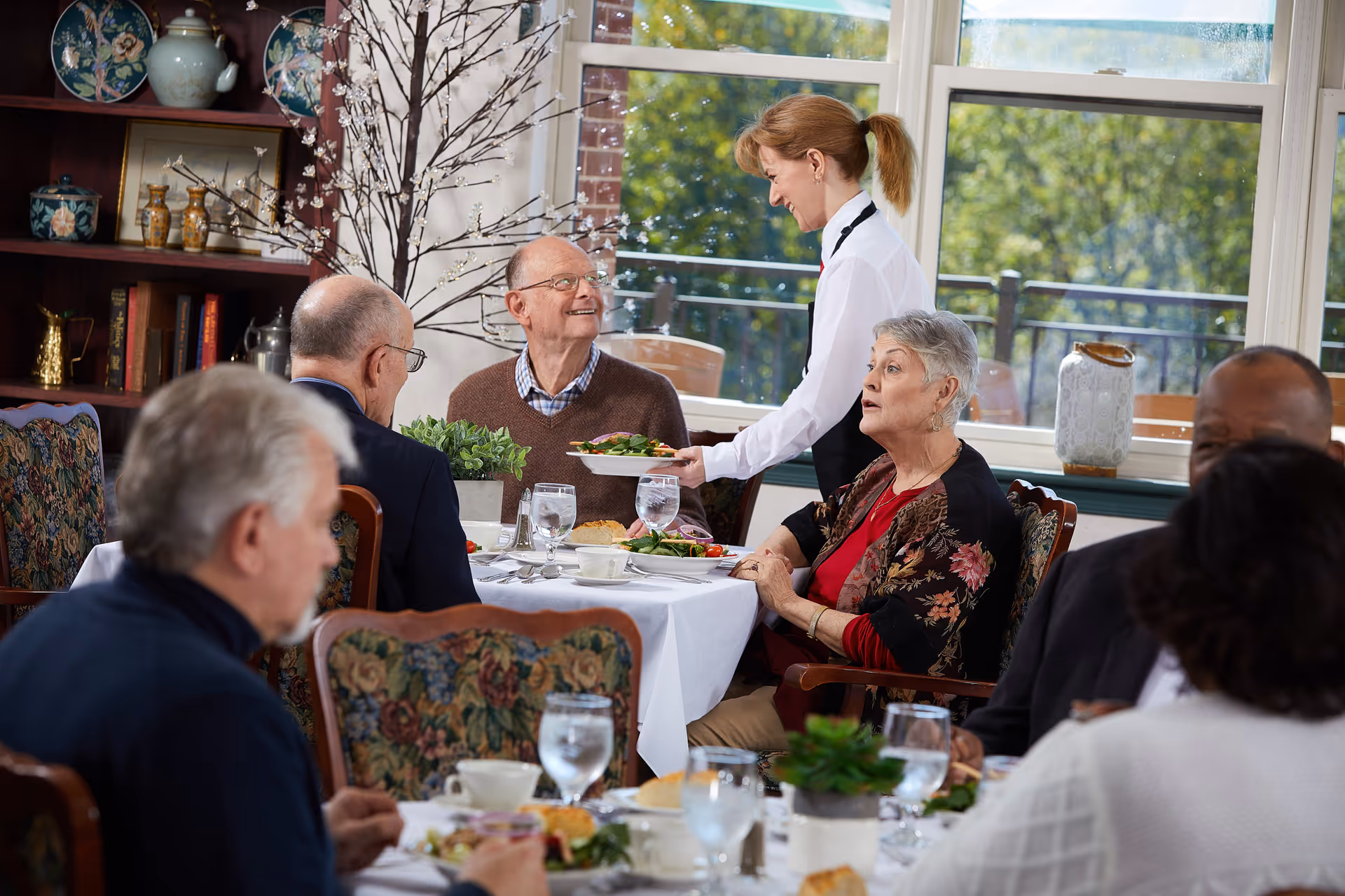 A group of elderly people seated around a dining table in a well-lit room with large windows. A waitress is serving a plate of food to a smiling elderly man. The table is set with plates, glasses of water, and some food items. The room has floral upholstered chairs and a wooden shelf with decorative items in the background.