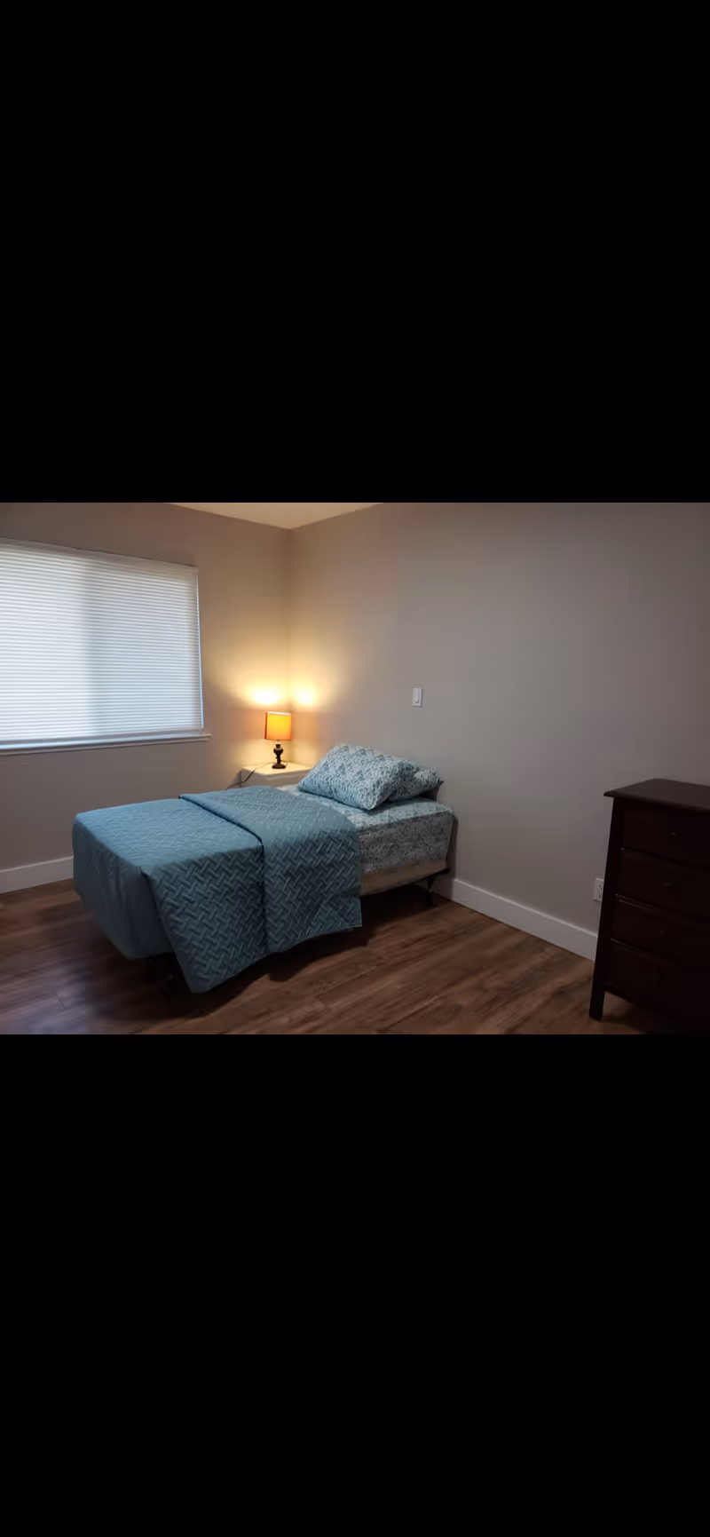 Simple bedroom with a twin bed dressed in teal bedding, a bedside table with a lit lamp, a window with blinds, and a dresser on a wood floor.
