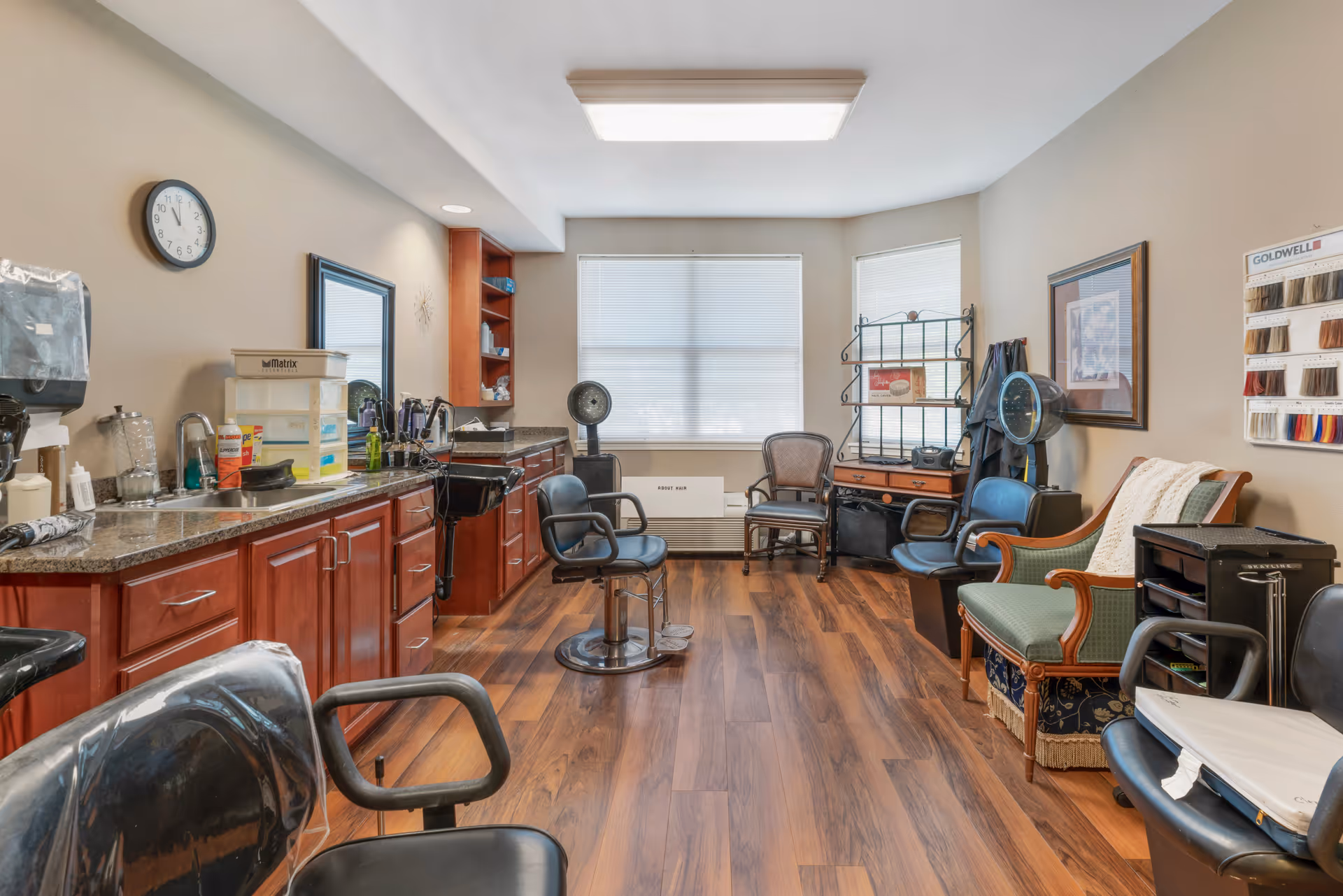 Interior view of a hair salon room with wooden flooring, multiple black salon chairs, a sink for washing hair, a large mirror, shelves with hair products, a clock on the wall, and a window with blinds letting in natural light. There is also a green upholstered bench, a rack with hair color samples, and hair drying equipment.