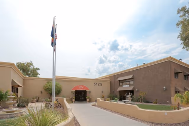 Exterior view of Bella Vita Health & Rehabilitation Center showing a beige building with the address number 5125 above the entrance. There are two flagpoles with flags, a bicycle parked near the entrance, and landscaped areas with plants and a small fountain.