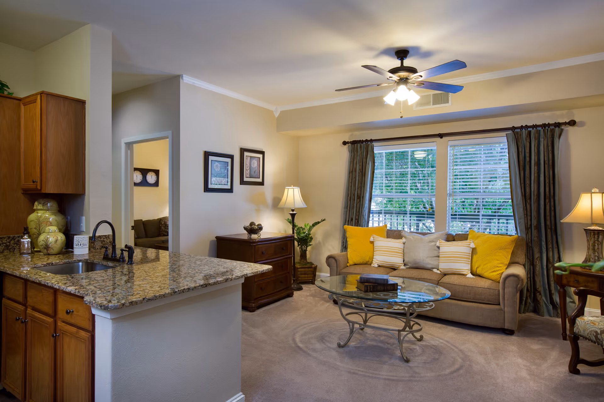A cozy living room area with a beige sofa adorned with yellow and striped pillows, a glass coffee table with books on it, a wooden side table with a lamp, and a ceiling fan with lights. The room has large windows with curtains letting in natural light. Adjacent to the living room is a kitchen area with granite countertops, wooden cabinets, and a sink with a faucet.