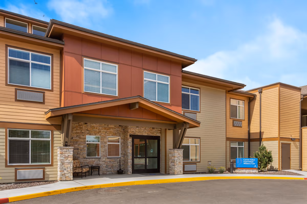 Exterior view of a two-story memory care facility building with beige and reddish-brown siding, multiple windows with blinds, a covered entrance supported by stone pillars, and a small seating area with two chairs and a table near the entrance. A blue sign near the entrance reads 'Carson Tahoe Expressions Memory Care'.