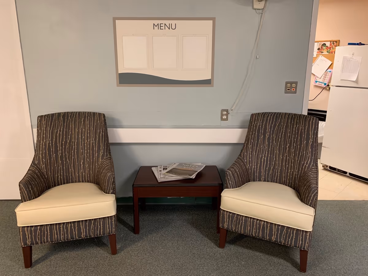 Two patterned armchairs with light-colored seat cushions are placed on either side of a small wooden table holding some newspapers. Behind the chairs is a light blue wall with a blank menu board mounted on it. To the right, there is a partial view of a white refrigerator and a bulletin board with papers pinned on it.