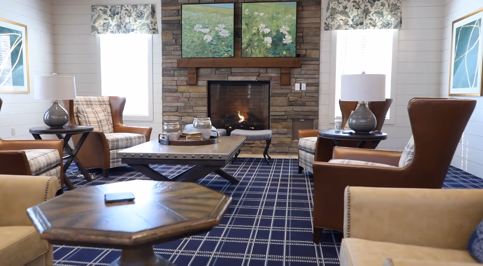 A cozy living room area with a stone fireplace in the center, flanked by two windows with floral valances. The room features four leather armchairs with plaid cushions arranged around a studded coffee table. There are side tables with lamps next to the chairs, and a patterned blue carpet covers the floor. Above the fireplace are two paintings of white flowers on a green background.