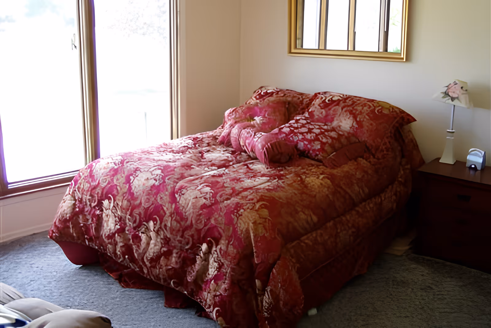 A bedroom with a bed covered in a red and pink patterned comforter and matching pillows. There is a wooden nightstand with a floral lamp and a small device on it. A large window with wooden trim is on the left side, and a framed mirror hangs on the wall above the bed.