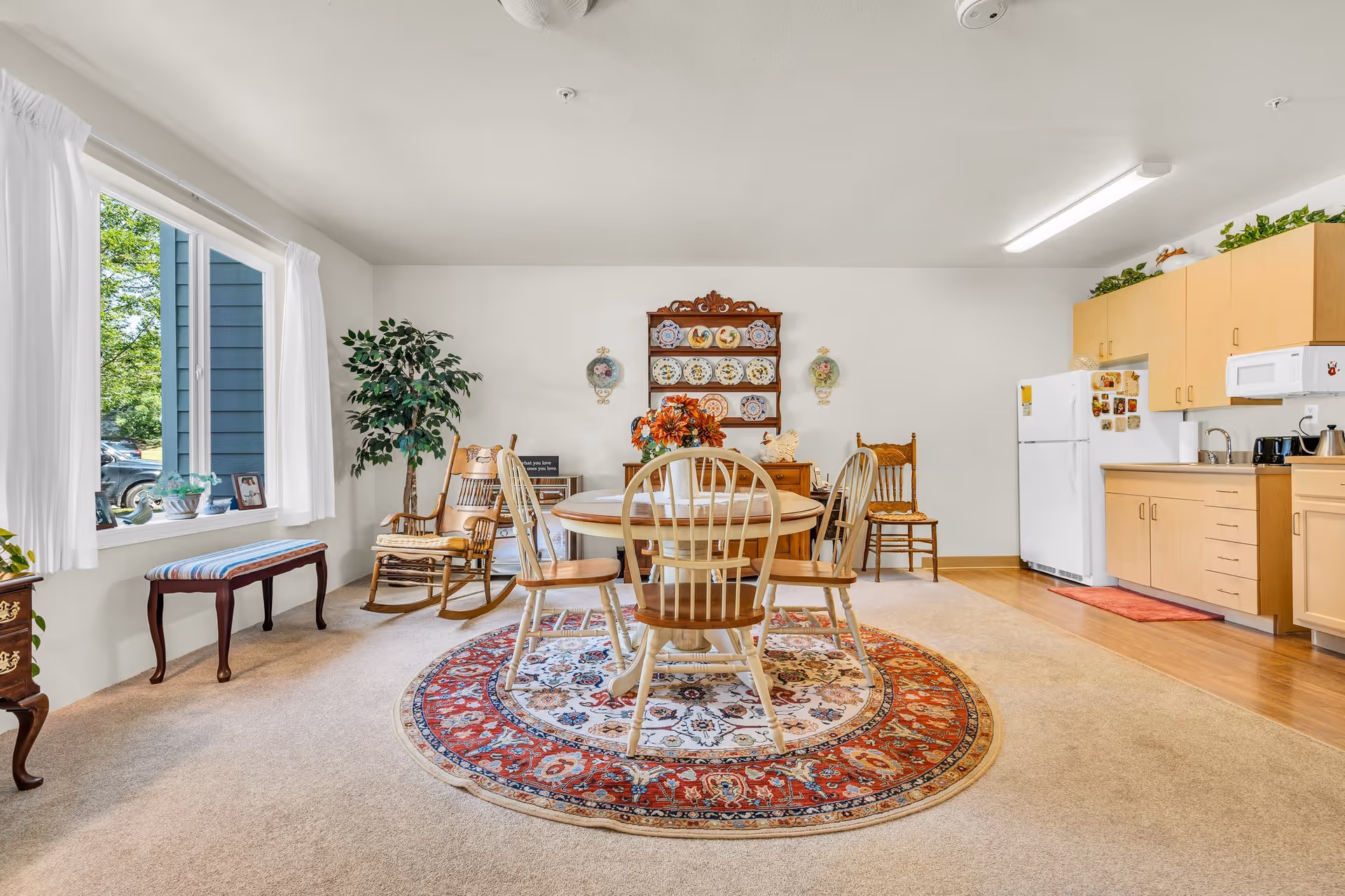 A bright and cozy senior living dining area with a round wooden table surrounded by six chairs on a decorative round rug. The room features a large window with white curtains, a rocking chair, a wooden bench, a potted plant, and a wooden hutch displaying decorative plates. The kitchen area with light wood cabinets, a white refrigerator, microwave, and coffee maker is visible in the background.