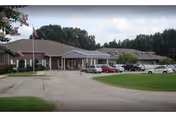 Front exterior of a single-story senior living facility with a covered entrance, flagpole, parked cars, and surrounding trees.