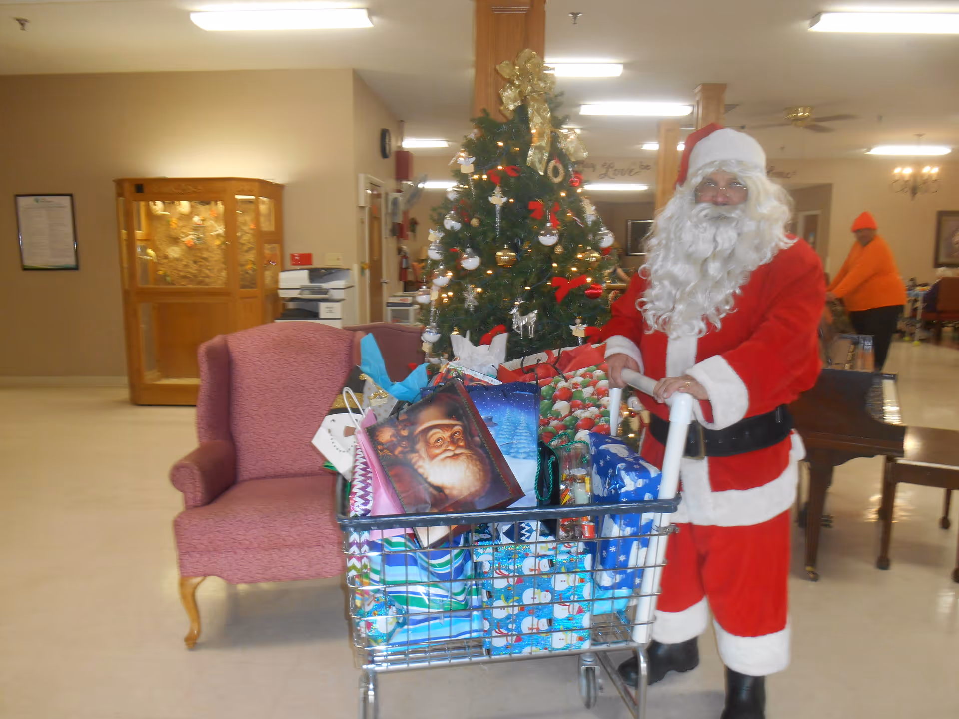 A person dressed as Santa Claus stands indoors next to a shopping cart filled with wrapped Christmas presents. Behind them is a decorated Christmas tree with ornaments and lights. The room has beige walls, a pink upholstered chair, a piano, and another person in an orange outfit in the background.