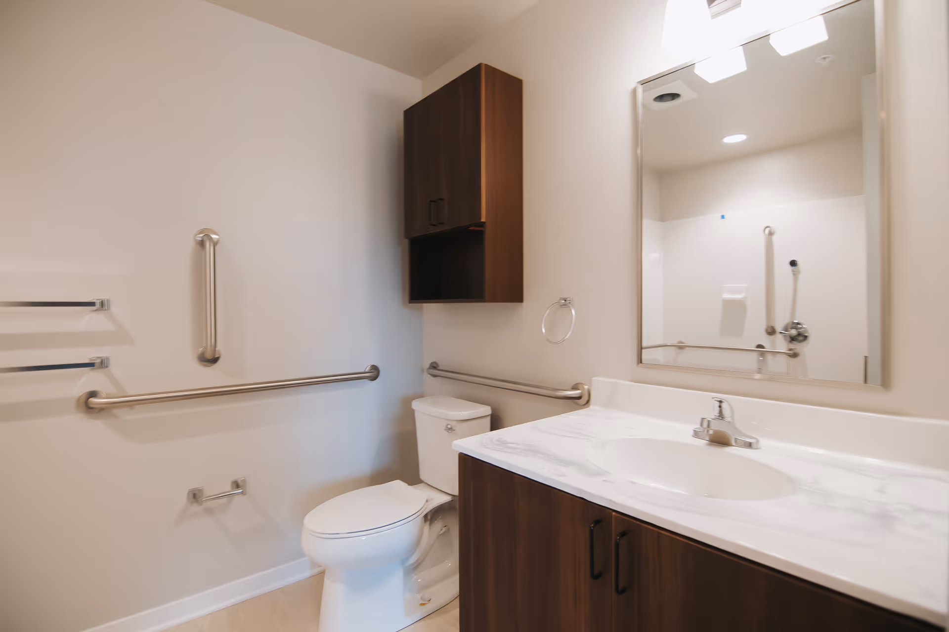 A clean and modern bathroom with a white toilet, a marble countertop sink with a wooden cabinet below, a large mirror above the sink, and a wooden wall cabinet. The bathroom has stainless steel grab bars on the walls for accessibility and a towel ring next to the sink.