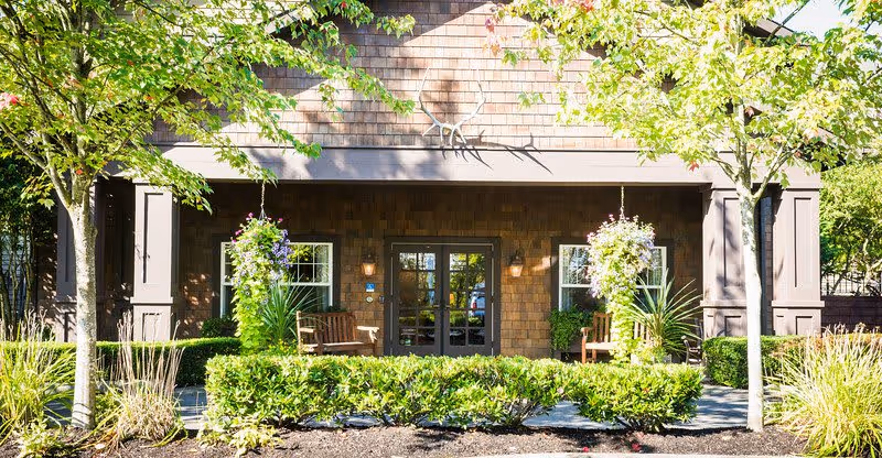 Front entrance of a building with brown shingle siding, two windows on either side of a double glass door, two wooden benches, hanging flower baskets, and neatly trimmed bushes and trees in the foreground.