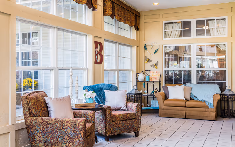 A cozy sitting area in a senior living facility with two patterned armchairs and a brown sofa, all adorned with light-colored pillows and blue throws. The space features large windows with decorative valances, a small round table with flowers, and wall decorations including a large letter B and butterfly ornaments. The floor is tiled and the room is well-lit with natural light.