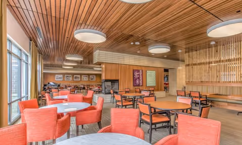 A spacious dining room with multiple round tables surrounded by orange upholstered chairs. The room features a wooden ceiling with circular light fixtures, large windows on the left side allowing natural light, and a fireplace in the back wall. The walls have wooden paneling and framed pictures, creating a warm and inviting atmosphere.