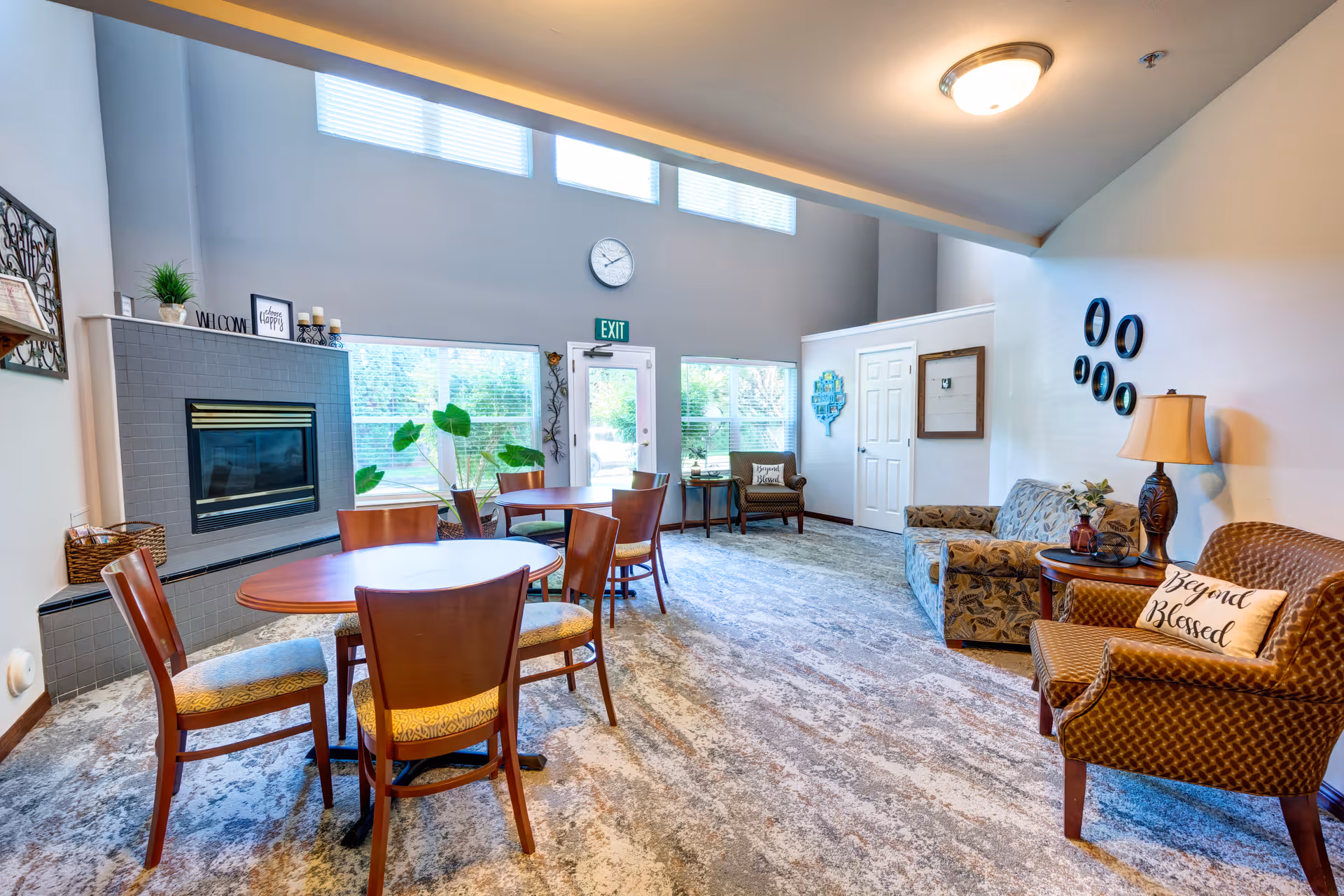 A bright and spacious common area in an assisted living facility featuring a fireplace with decorative items on the mantel, round wooden tables with chairs, comfortable armchairs with cushions, a side table with a lamp, large windows letting in natural light, and a carpeted floor.